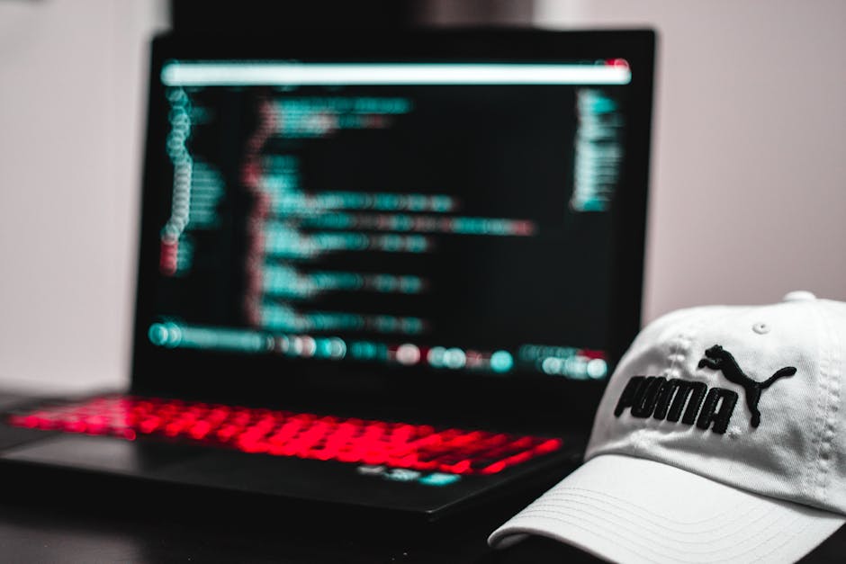 This image features a close-up view of a laptop with a black keyboard illuminated by red backlighting, displaying lines of code on its screen with a blurred background. In the foreground, a white Puma