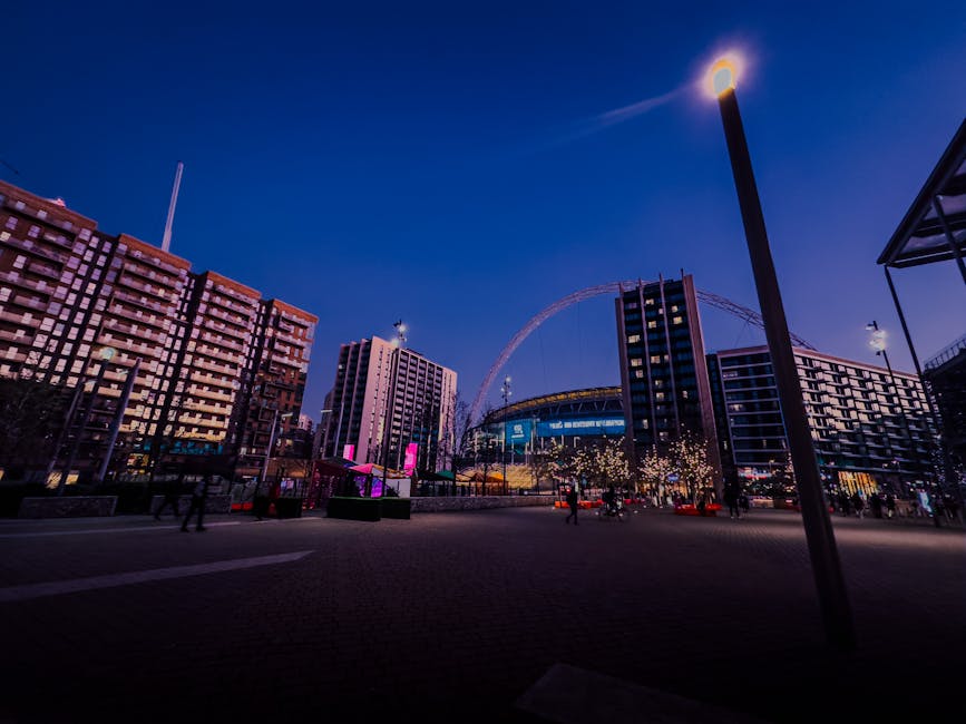 A wide view of Wembley Park at dusk featuring modern multi-storey buildings with illuminated windows and outdoor lighting, including a tall street lamp in the foreground casting a glow. The sky is a d
